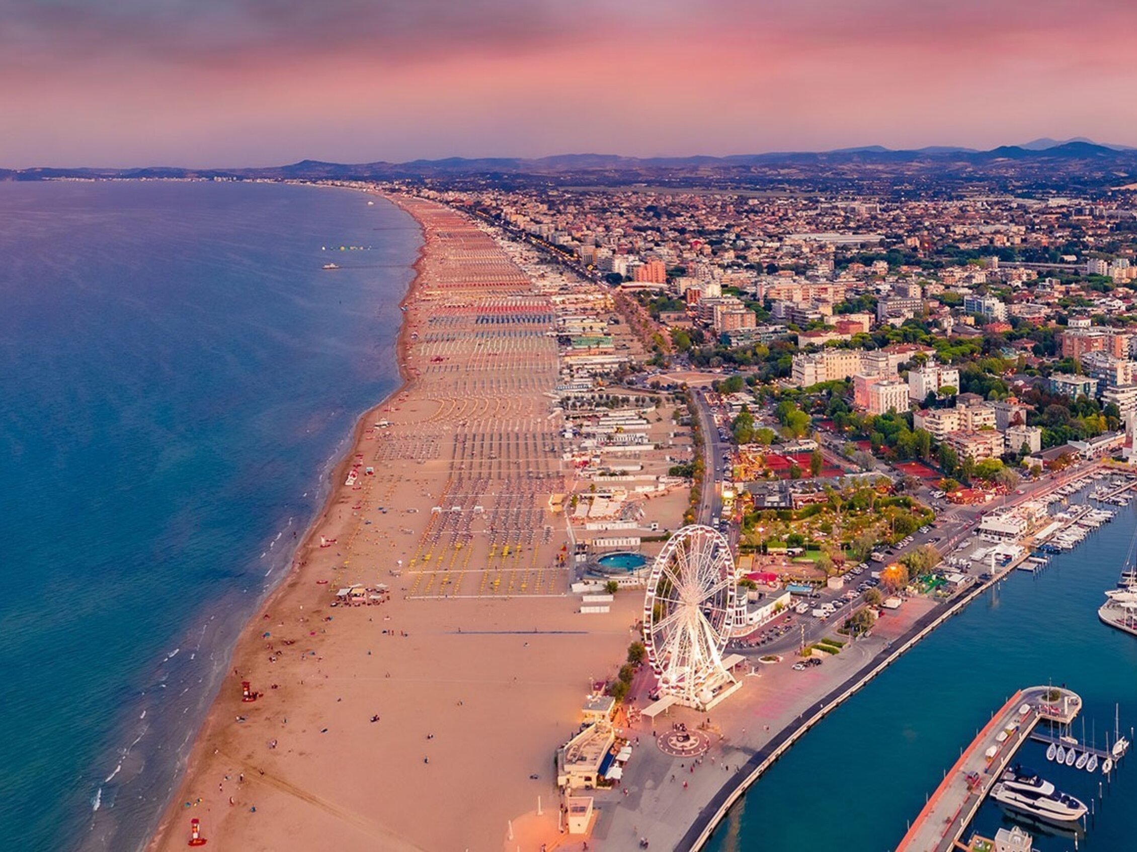 Aerial view of Rimini’s coastline at sunset, featuring the long golden beach, marina, Ferris wheel, and the city stretching from the sea towards the hills in the background.﻿