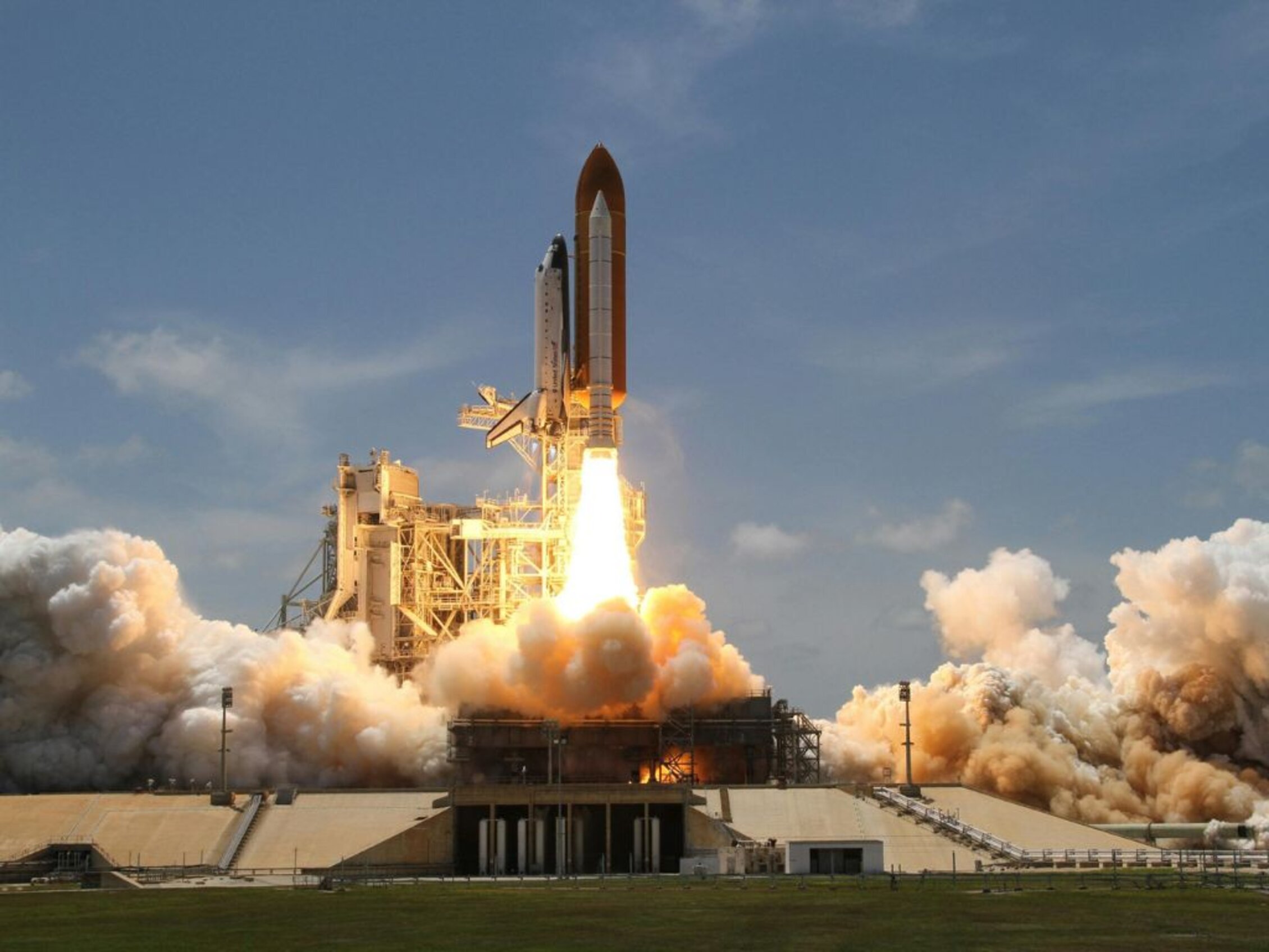 Space rocket liftoff with bright orange flames and thick white smoke trails against a clear sky
