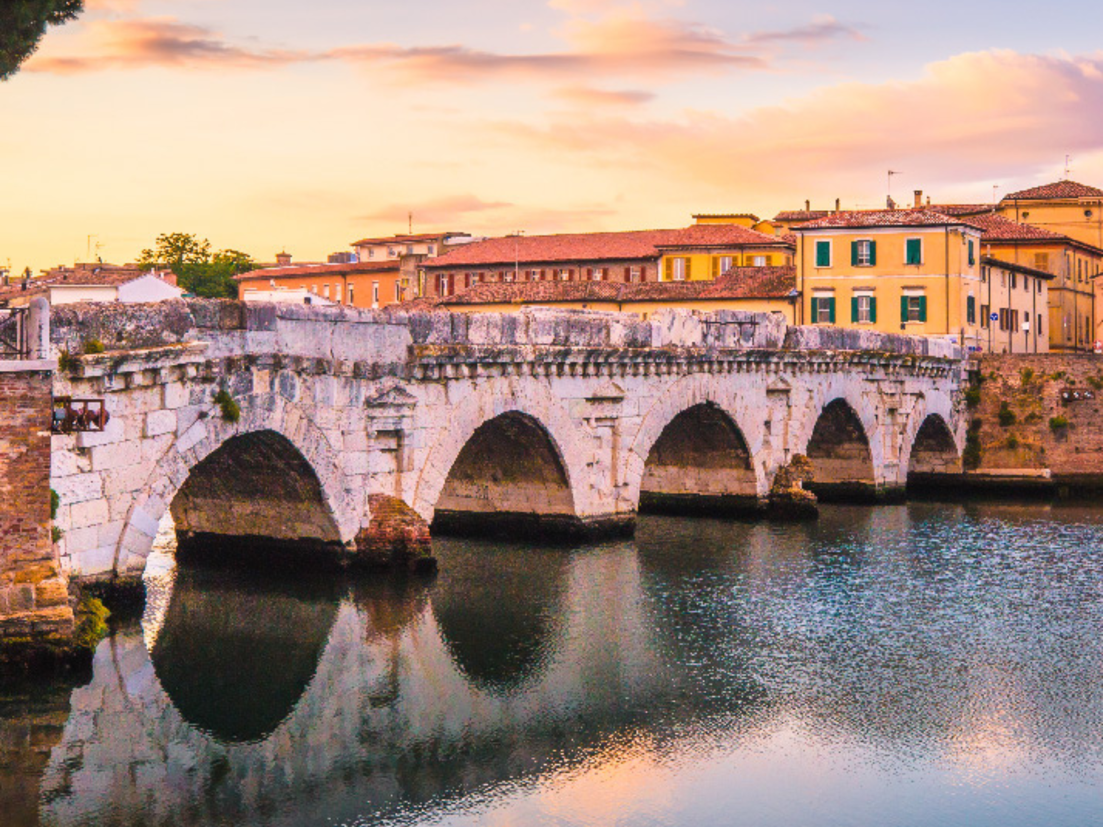 Ancient Roman Tiberius Bridge (Ponte di Tiberio) in Rimini during golden hour, reflecting in the Marecchia River waters.